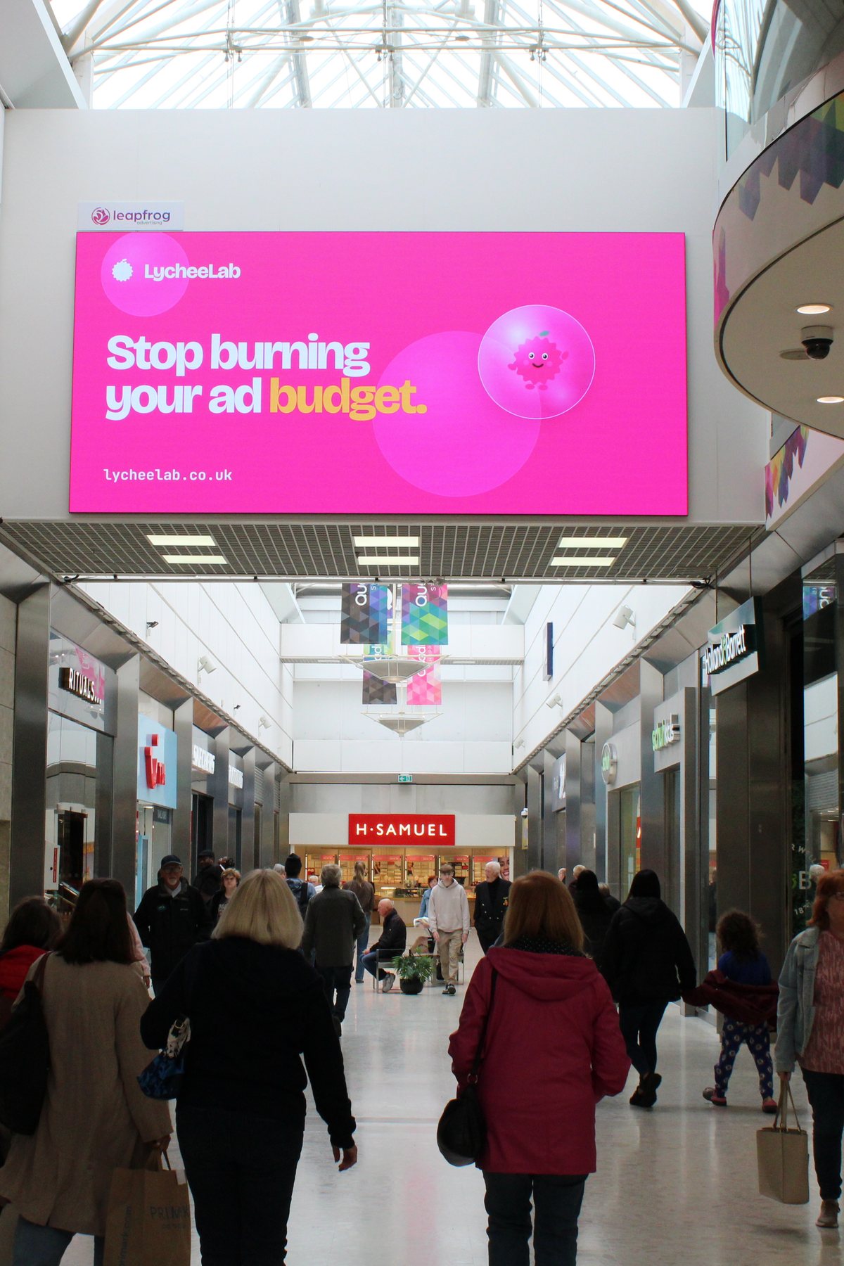 The Lychee Lab billboard on a shopping centre screen: Stop burning your ad budget, lycheelab.co.uk.